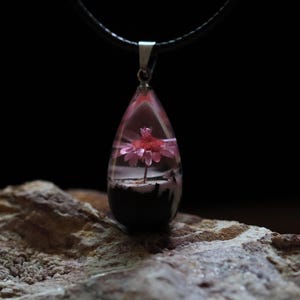 Può includere: Collana con pendente a goccia con un fiore rosa racchiuso in resina trasparente. Il pendente è appeso a un cordino nero con chiusura color argento. Lo sfondo è scuro e il pendente poggia su una superficie di pietra strutturata.