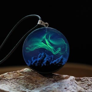 Può includere: Collana con pendente rotondo e cordino nero. Il pendente presenta un design blu e verde che ricorda l'aurora boreale sopra una catena montuosa. Il pendente è posto su uno sfondo scuro.