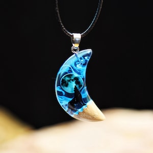 Può includere: Ciondolo a forma di luna crescente con cordino nero. Il ciondolo presenta un design in resina blu con la sagoma di un lupo che ulula alla luna piena. La parte inferiore della luna è di colore legno chiaro.