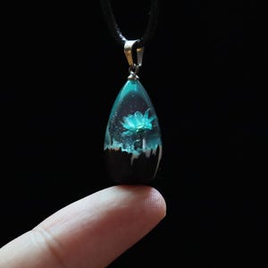 Può includere: Una collana con pendente a goccia con cordino nero. Il pendente è trasparente con un fiore color verde acqua racchiuso all'interno. La parte inferiore del pendente è marrone scuro con accenti bianchi. Il pendente è tenuto da un'anello color argento.