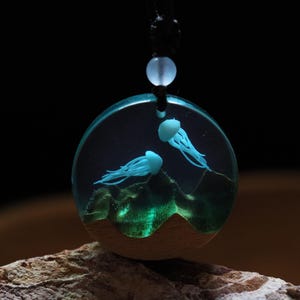 Può includere: Una collana con pendente rotondo con due meduse blu all'interno di un cerchio trasparente pieno di resina. Il pendente ha una base in legno ed è appeso a un cordino nero. Le meduse sono su uno sfondo scuro con accenti verdi.