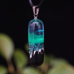 Può includere: Un ciondolo artigianale con un design unico. Il ciondolo in resina presenta strati di blu, verde e nero, creando un effetto visivo sorprendente. È appeso a un cordino nero con un fermaglio color argento. Lo sfondo è scuro.