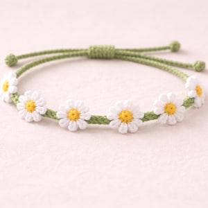May include: A daisy chain bracelet featuring white crocheted daisy flowers with yellow centers. The flowers are attached to a green crocheted band. The bracelet has an adjustable green cord closure.