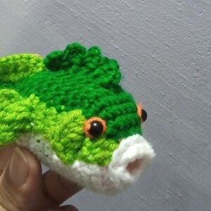 May include: A small, handcrafted crochet fish toy featuring a vibrant green and lime green textured body, a white underbelly, and a white mouth. The fish has two prominent orange eyes with black pupils and detailed fins along the top and sides.
