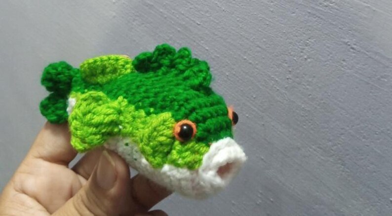 May include: A small, handcrafted crochet fish toy featuring a vibrant green and lime green textured body, a white underbelly, and a white mouth. The fish has two prominent orange eyes with black pupils and detailed fins along the top and sides.