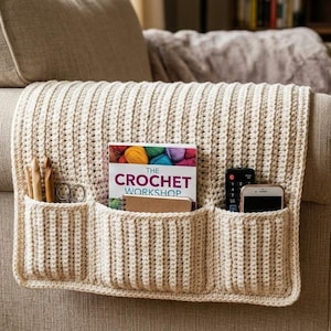 May include: Cream-colored, crocheted sofa armrest organizer with three pockets. The pockets hold a book titled "The Crochet Workshop", a remote control, a smartphone, crochet hooks, and scissors. The organizer is draped over a beige sofa arm.