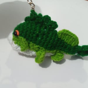 May include: A crocheted keychain shaped like a fish with a dark green back, a light green tail and fins, and a white underbelly. The fish features a single visible orange eye and is attached to a small metal chain and ring for hanging.