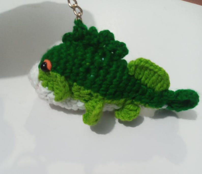 May include: A crocheted keychain shaped like a fish with a dark green back, a light green tail and fins, and a white underbelly. The fish features a single visible orange eye and is attached to a small metal chain and ring for hanging.