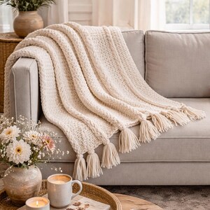 May include: Cream-colored, chunky knit throw blanket with tassels draped over a gray sofa. The blanket has a textured pattern and a soft, inviting appearance. The tassels hang from the edge of the blanket. The scene includes a coffee table with a tray, candles, and a mug.