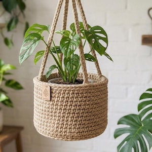 May include: A woven, tan-coloured hanging plant basket, holding a green plant with large, patterned leaves. The basket is suspended by thick, woven rope. A small wooden tag with a leaf symbol is attached to the basket.