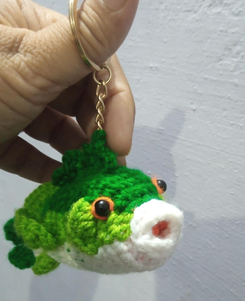 May include: A crocheted fish keychain featuring a vibrant green body with a white underbelly, orange-rimmed black eyes, and a small red mouth. The plush fish is attached to a gold-toned metal split ring and a short chain for easy attachment.