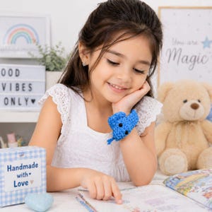 Puede incluir: Una niña pequeña lleva una pulsera azul en forma de corazón hecha a ganchillo. La pulsera está en su muñeca. La niña sonríe y mira un libro. Un oso de peluche y una caja con el texto "Handmade with Love" están en el fondo.