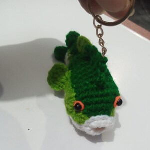 May include: A crocheted fish keychain featuring dark green and light green yarn, a white mouth area, and two black bead eyes with orange yarn accents. The fish is attached to a gold-toned metal split ring by a short chain, allowing for easy attachment.