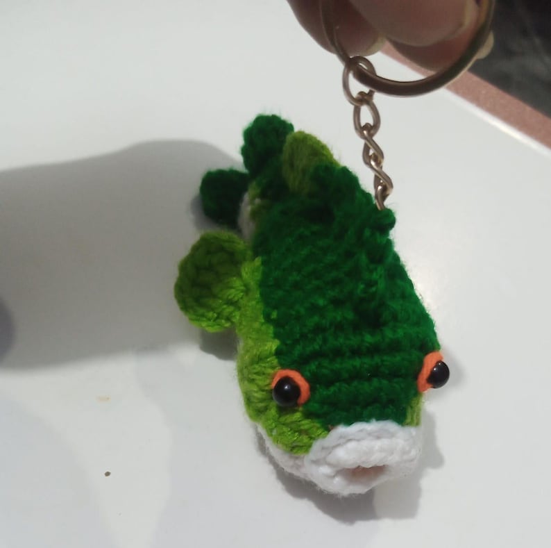 May include: A crocheted fish keychain featuring dark green and light green yarn, a white mouth area, and two black bead eyes with orange yarn accents. The fish is attached to a gold-toned metal split ring by a short chain, allowing for easy attachment.