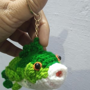May include: A crocheted fish keychain featuring a vibrant green body with a white underbelly, orange-rimmed black eyes, and a small red mouth. The plush fish is attached to a gold-toned metal split ring and a short chain for easy attachment.