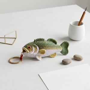 May include: A crocheted fish keychain in shades of green, white, and yellow, with an orange eye and a gold keyring. The fish is lying on a white surface. The keychain is a unique accessory.