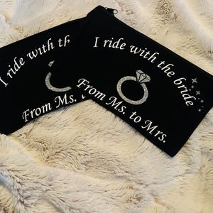 May include: Two black zippered pouches with white text that reads "I ride with the bride From Ms. to Mrs." and a glittery ring design. The pouches are on a fluffy, cream-colored surface.