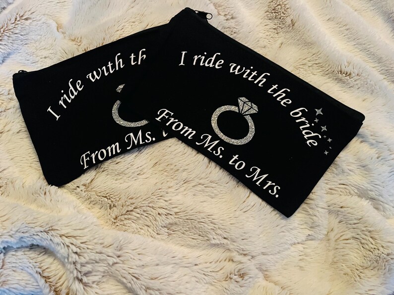 May include: Two black zippered pouches with white text that reads "I ride with the bride From Ms. to Mrs." and a glittery ring design. The pouches are on a fluffy, cream-colored surface.