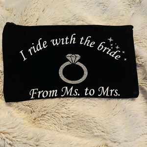 May include: Black zippered pouch with white text that reads "I ride with the bride" and "From Ms. to Mrs." A silver glitter ring graphic is in the center. The pouch is rectangular and sits on a fluffy, cream-colored surface.