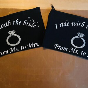 May include: Two black zippered pouches with the text "I ride with the bride" and "From Ms. to Mrs." in white script. A silver ring graphic is centered below the text. The pouches are on a wooden surface.