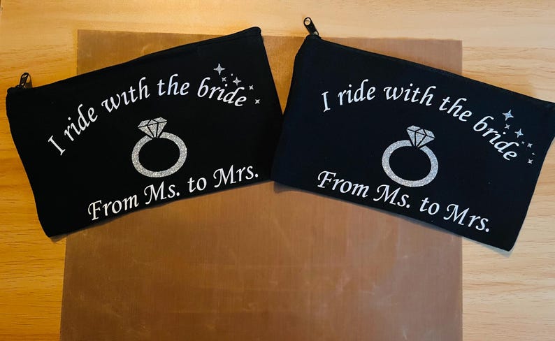 May include: Two black zippered pouches with the text "I ride with the bride" and "From Ms. to Mrs." in white script. A silver ring graphic is centered below the text. The pouches are on a wooden surface.