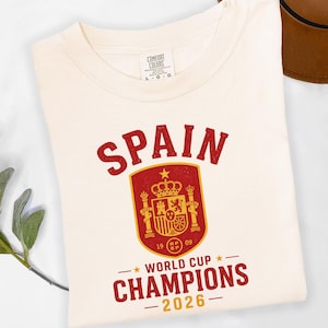 May include: A cream-colored t-shirt featuring a red and gold graphic. The text reads SPAIN at the top, with a central shield emblem containing a crown and crest. Below the shield, the text reads WORLD CUP CHAMPIONS 2026 in red and gold lettering.