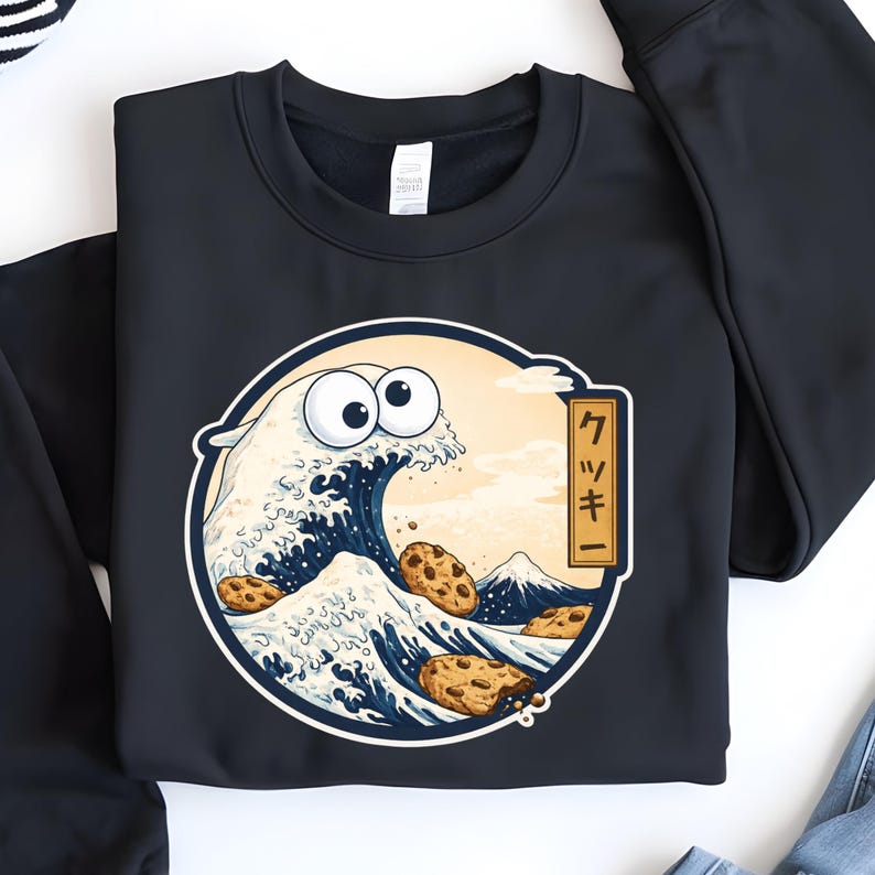 Pode incluir: Camisola preta com um design de onda de desenho animado. O design inclui um personagem com olhos de biscoito, biscoitos e uma montanha, emoldurados em um c&iacute;rculo com texto japon&ecirc;s.