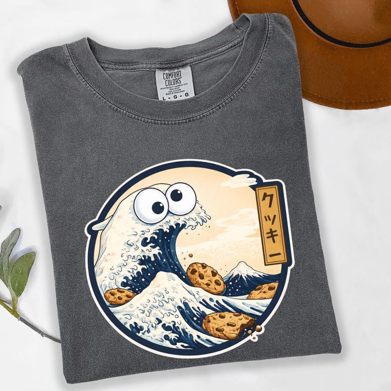Pode incluir: Camiseta cinza Comfort Colors com um gr&aacute;fico circular de uma onda com olhos de biscoito, biscoitos com gotas de chocolate e uma paisagem montanhosa. O gr&aacute;fico inclui texto japon&ecirc;s. A camiseta tem gola redonda.