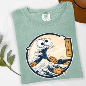 Pode incluir: Uma camiseta verde claro com um gr&aacute;fico circular que apresenta uma onda com olhos arregalados e biscoitos com gotas de chocolate. O design &eacute; inspirado na obra de arte japonesa, "A Grande Onda de Kanagawa". O texto japon&ecirc;s diz "Cookie".