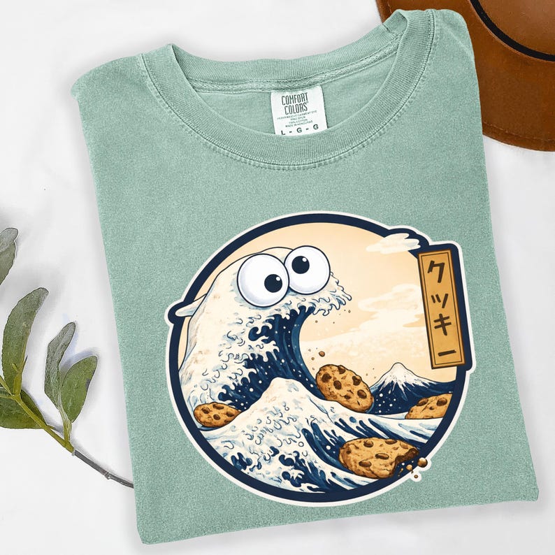 Pode incluir: Uma camiseta verde claro com um gr&aacute;fico circular que apresenta uma onda com olhos arregalados e biscoitos com gotas de chocolate. O design &eacute; inspirado na obra de arte japonesa, "A Grande Onda de Kanagawa". O texto japon&ecirc;s diz "Cookie".