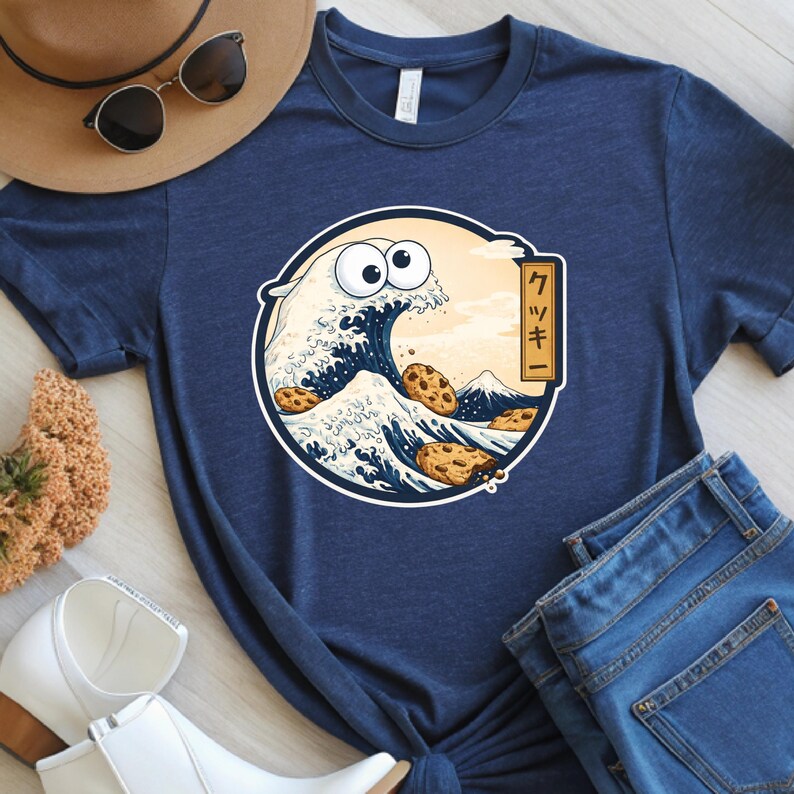 Pode incluir: Camiseta azul marinho com um desenho de biscoito de desenho animado em uma onda, com o texto japon&ecirc;s "クッキー" (kukkī). O design inclui uma montanha e biscoitos na onda. A camiseta &eacute; combinada com jeans azuis, um chap&eacute;u e &oacute;culos de sol.