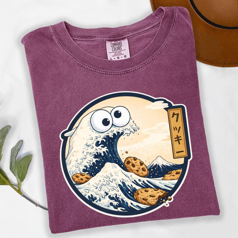 Pode incluir: Camiseta Comfort Colors bord&ocirc; com um gr&aacute;fico circular. O gr&aacute;fico retrata uma onda com olhos arregalados e biscoitos com gotas de chocolate, inspirada na arte japonesa. Texto japon&ecirc;s &agrave; direita.
