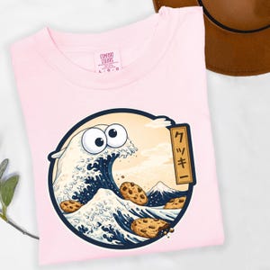 Pode incluir: Camiseta rosa claro Comfort Colors com um gr&aacute;fico circular. O gr&aacute;fico retrata uma onda com olhos arregalados e biscoitos com pepitas de chocolate, inspirada na arte japonesa. O design inclui uma montanha e texto japon&ecirc;s.