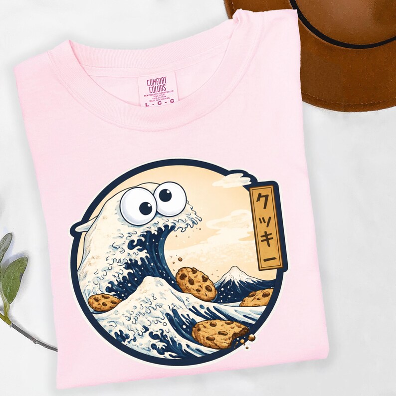 Pode incluir: Camiseta rosa claro Comfort Colors com um gr&aacute;fico circular. O gr&aacute;fico retrata uma onda com olhos arregalados e biscoitos com pepitas de chocolate, inspirada na arte japonesa. O design inclui uma montanha e texto japon&ecirc;s.