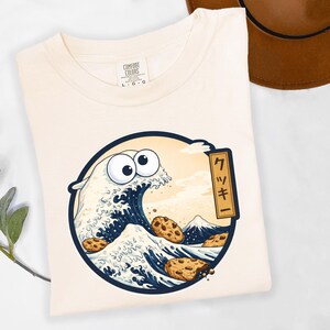 Pode incluir: Camiseta creme com um desenho de onda de desenho animado, com olhos arregalados e biscoitos com pepitas de chocolate. O desenho est&aacute; dentro de um c&iacute;rculo azul marinho, com texto japon&ecirc;s e uma montanha no fundo. A camisa tem uma etiqueta Comfort Colors.