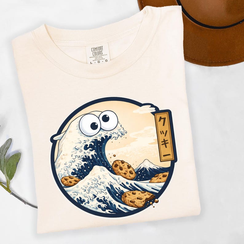 Pode incluir: Camiseta creme com um desenho de onda de desenho animado, com olhos arregalados e biscoitos com pepitas de chocolate. O desenho est&aacute; dentro de um c&iacute;rculo azul marinho, com texto japon&ecirc;s e uma montanha no fundo. A camisa tem uma etiqueta Comfort Colors.
