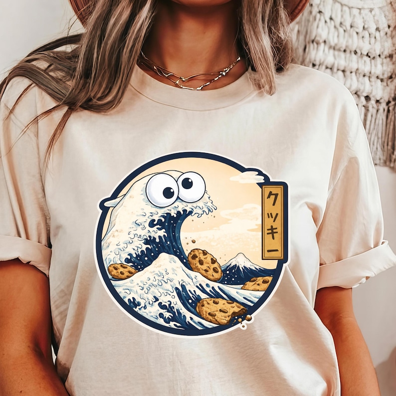 Pode incluir: Camiseta bege com um gr&aacute;fico circular que retrata uma onda com olhos arregalados e biscoitos com pepitas de chocolate. O design &eacute; inspirado na arte japonesa, com uma montanha e texto em uma moldura retangular. O texto diz "クッキー".
