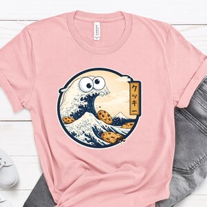 Pode incluir: T-shirt rosa claro com um gr&aacute;fico circular que retrata uma onda com ondas em forma de biscoito e um rosto de desenho animado. O gr&aacute;fico inclui a palavra japonesa para "biscoito". A camisa &eacute; feita de um material macio.