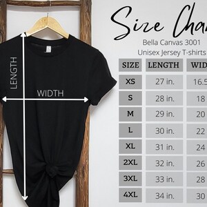 Pode incluir: Uma camiseta unissex preta Bella Canvas 3001 pendurada em uma escada de madeira. A imagem inclui uma tabela de tamanhos com medidas em polegadas para os tamanhos XS a 4XL. O gr&aacute;fico lista o comprimento e a largura para cada tamanho.