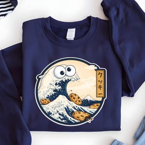 Pode incluir: Camisola azul marinho com um design de desenho animado. O design apresenta uma onda com olhos arregalados e biscoitos com pepitas de chocolate, inspirada em "A Grande Onda de Kanagawa". O texto japon&ecirc;s diz "Cookie".