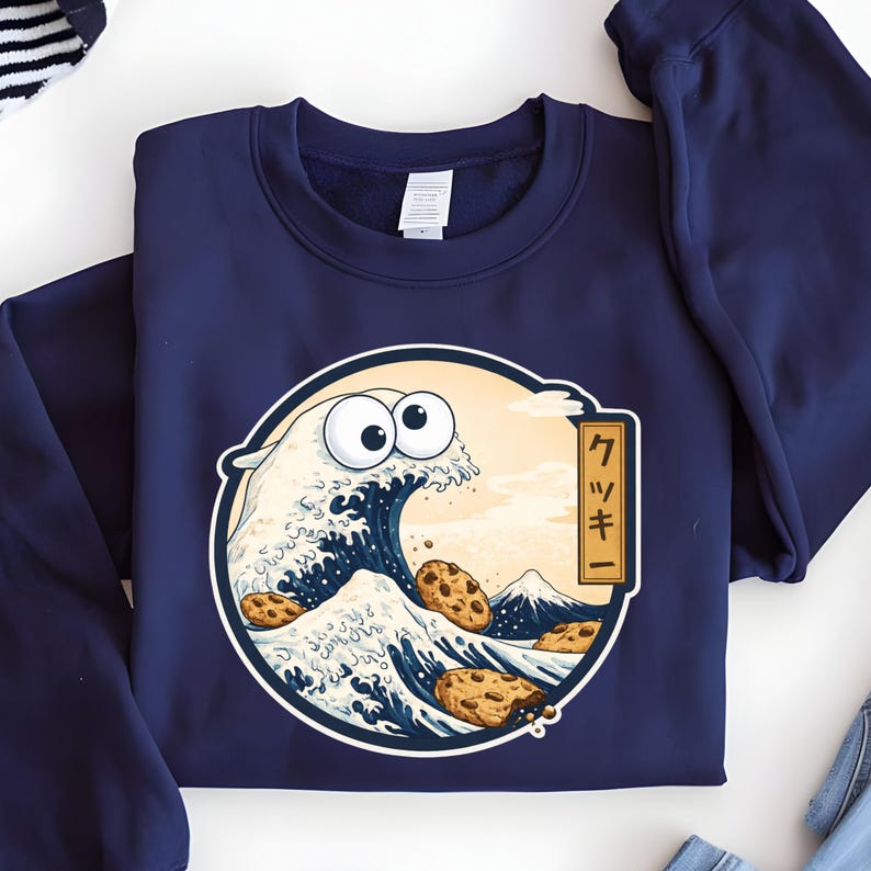 Pode incluir: Camisola azul marinho com um design de desenho animado. O design apresenta uma onda com olhos arregalados e biscoitos com pepitas de chocolate, inspirada em "A Grande Onda de Kanagawa". O texto japon&ecirc;s diz "Cookie".