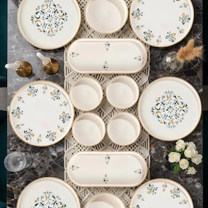 Op de afbeelding: Een 14-delige servies set met witte borden, kommen en serveerschalen. De borden hebben een gouden rand en een centraal bloemmotief in groen, geel en blauw. De set is op een decoratieve tafelsetting geplaatst.