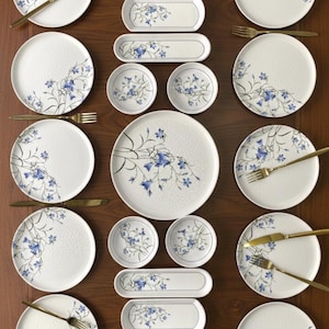 May include: A white dinner service featuring a blue and green floral pattern. The set includes plates, bowls, and serving dishes, accompanied by gold-coloured cutlery. The arrangement is displayed on a dark brown wooden surface.