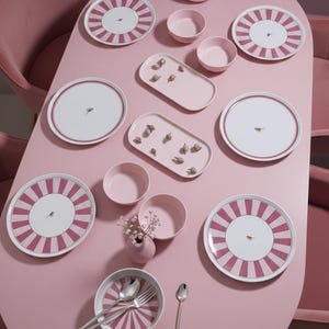 Peut inclure: Une table rose avec une nappe blanche ornée d'un motif rayé rose et d'une petite illustration d'oiseau. L'ensemble comprend des bols, des plats de service et des couverts. Un petit vase avec des fleurs est également présent.