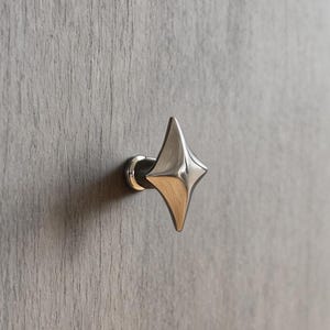 Op de afbeelding: Een zilveren, stervormige kastknop op een grijze houten lade. De knop heeft een vierpuntig sterontwerp en een gepolijste afwerking. De houtnerf is zichtbaar.