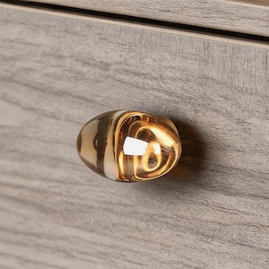 May include: Close-up of a cabinet drawer with a decorative, oval-shaped glass knob. The knob features a swirl design with amber and clear tones. The drawer is made of wood with a gray, textured finish.