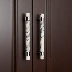 May include: Two cabinet pulls with a white and gray marble design. Each pull has a cylindrical shape with silver-colored metal accents. The pulls are mounted on a dark brown cabinet door.