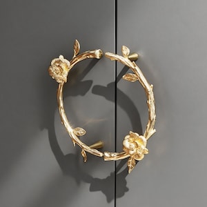 May include: Gold-colored cabinet door handles in a floral design. The handles are shaped like a circle with a rose at the top and bottom, and leaves and branches throughout. The handles are mounted on a gray cabinet door.