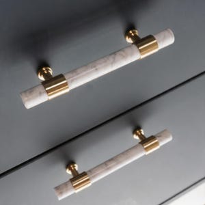 May include: Two cabinet pulls with a marble-like appearance and gold-colored accents. The cylindrical handles are white with gray veining, and are held in place by gold-colored rings and circular bases. The pulls are mounted on a dark gray surface.