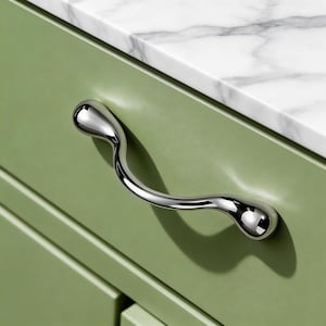 Könnte beinhalten: Ein polierter Chrom-Schrankgriff mit einem einzigartigen, geschwungenen Design. Der Griff ist an einer hellgrünen Schranktür befestigt, mit einer weißen Marmor-Arbeitsplatte im Hintergrund. Der Griff hat eine glatte, reflektierende Oberfläche.